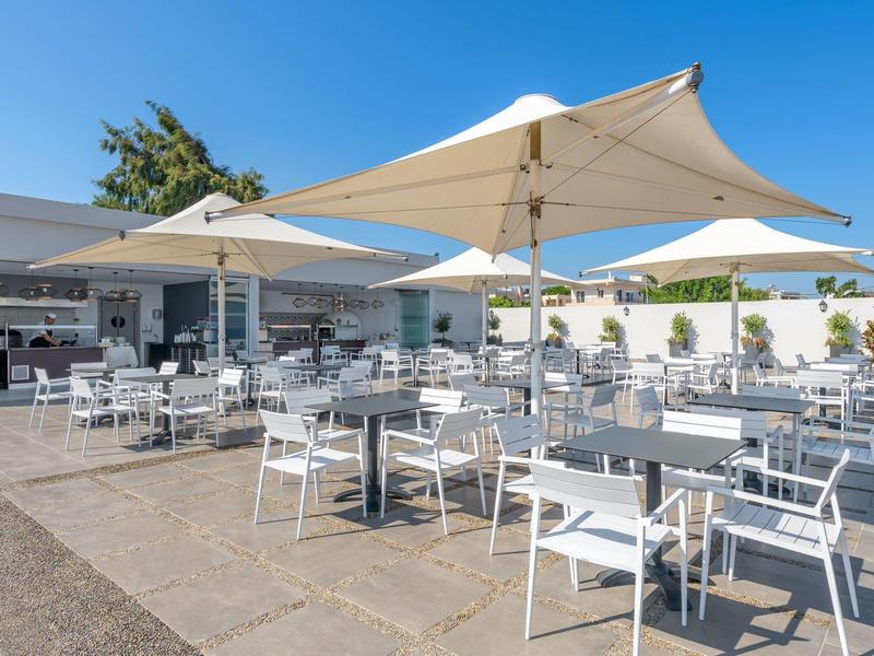 Terrasse mit weißen Tischen und Stühlen unter großen Sonnenschirmen bei klarem Himmel.