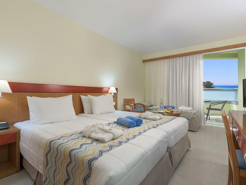 Helles Hotelzimmer mit zwei Betten, Tisch, Klimaanlage und Balkon mit Meerblick.