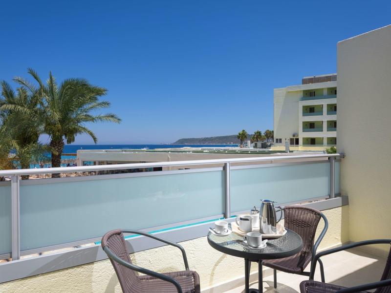 Balkon mit Glasgeländer, kleinem Tisch, zwei Stühlen, Palmen und Meer im Hintergrund bei blauem Himmel.