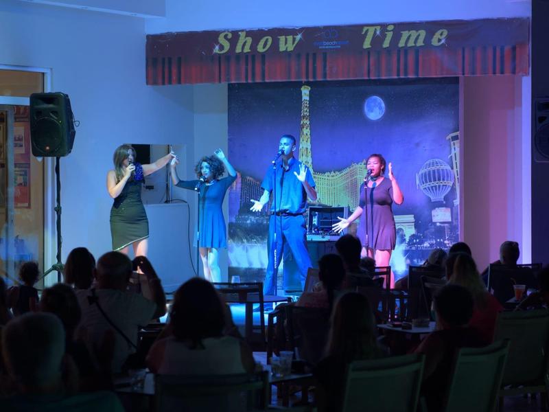 Bühne mit vier Sängern und Publikum in dunklem Saal, beleuchtet von blauem Licht unter Banner "Show Time".