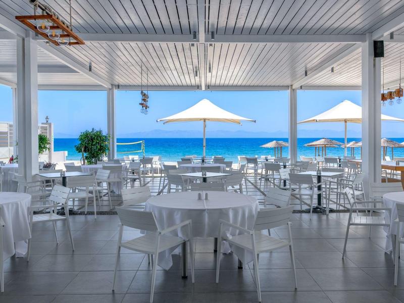 Freiluftrestaurant mit weißen Tischen und Stühlen, Blick auf Meer und Sonnenschirme.