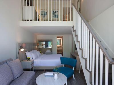 Modern furnished hotel room with double bed, sofa, blue chair, and staircase to the upper floor.