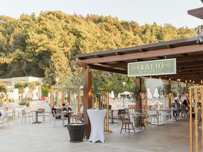 Offene Terrasse eines Cafés mit Tischen, Stühlen und Blick auf bewaldeten Hügel im Hintergrund.