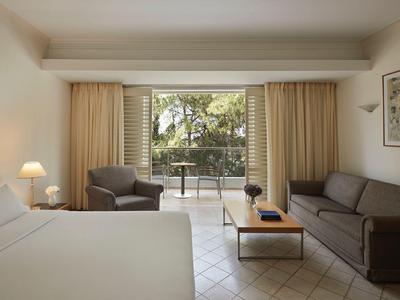 Hotel room with double bed, sofa, armchair, table, and balcony overlooking garden