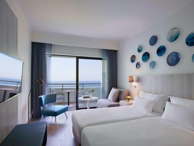 Modern hotel room with two beds, seating area, and sea view through large windows.