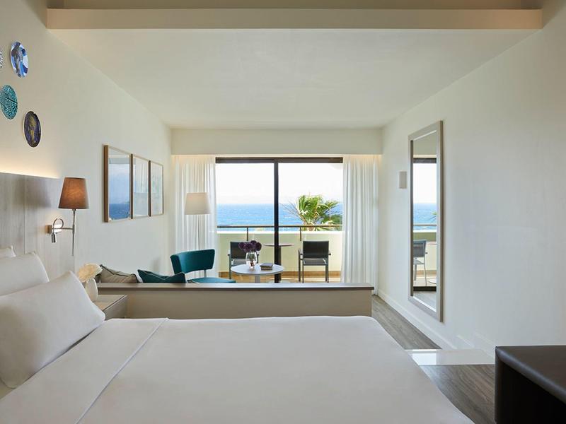 Modern hotel room with large bed, seating area, and sea view through glass doors.