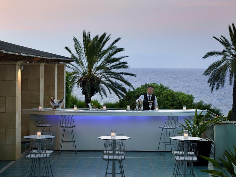 Modern outdoor bar area with white chairs, bar counter, and palm trees by the sea