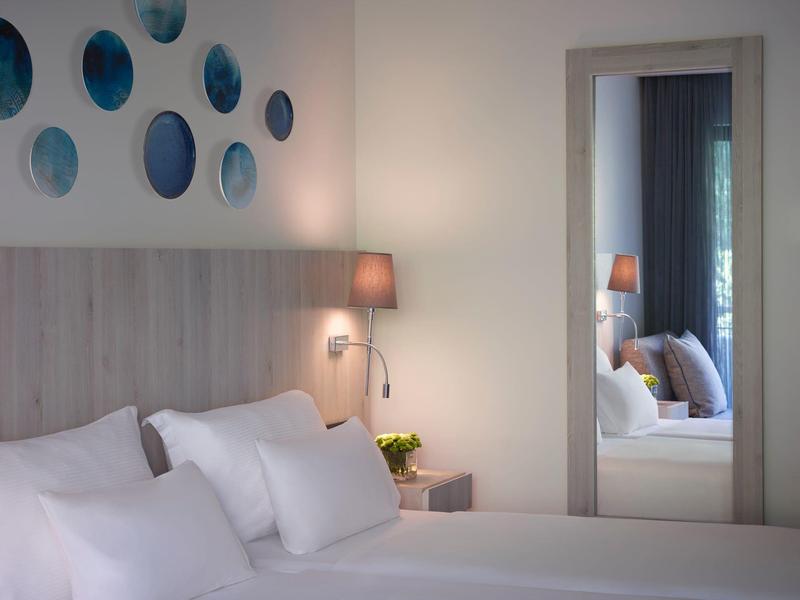Bright hotel room with a bed, multiple pillows, bedside table with lamp, and large mirror.