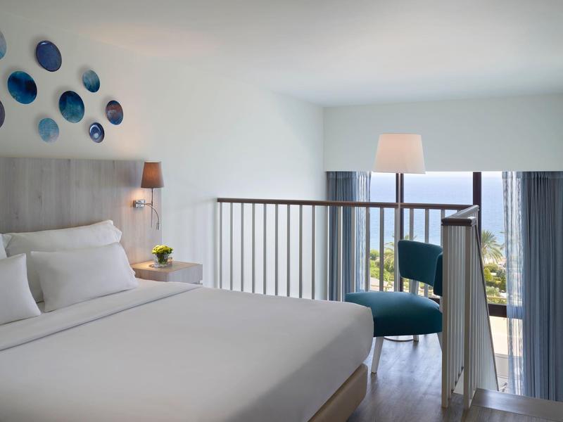 Bright hotel room with double bed, wall decoration, and lounge area with sea view.