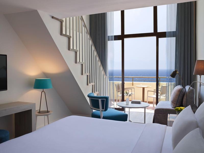 Modern hotel room with sea view, large bed, and seating area by floor-to-ceiling window.