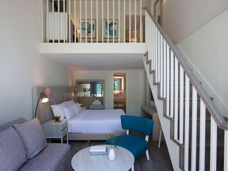 Modern furnished hotel room with double bed, sofa, blue chair, and staircase to the upper floor.