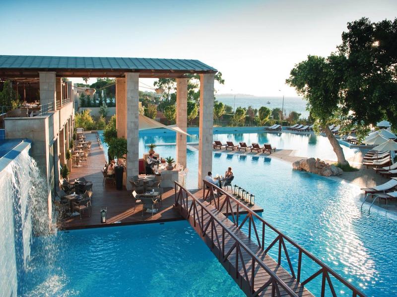 Luxury hotel pool with waterfall, wooden bridge, and sun loungers by the sea