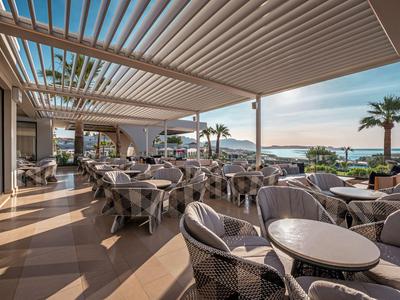 Überdachte Terrasse eines Hotels mit runden Tischen und Korbsesseln, Blick aufs Meer und Palmen.