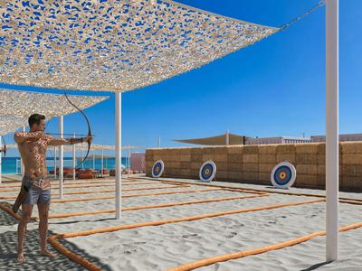 Zwei Personen üben Bogenschießen auf einem Sandplatz mit Zielscheiben bei klarem Himmel.