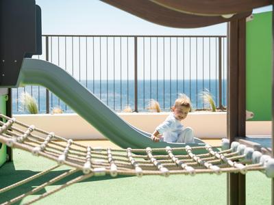 Kind spielt auf einem Spielplatz mit Rutsche, Balkon und Meerblick im Hintergrund.