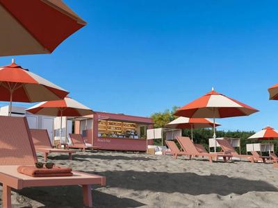 Strand mit Sonnenliegen und roten Sonnenschirmen vor klarem blauem Himmel, Hotel im Hintergrund.