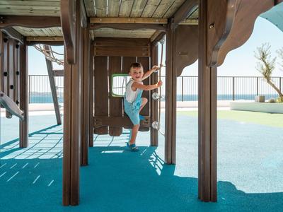 Kind spielt auf einem Holzspielplatz mit Blick aufs Meer bei sonnigem Wetter.