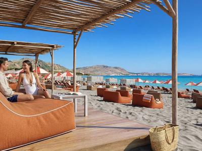 Ein Strand mit Sonnenschirmen und Sitzgelegenheiten unter klarem blauem Himmel neben zwei Personen auf einer überdachten Lounge.