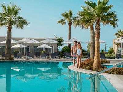 Ein Paar steht am Beckenrand eines blauen Pools mit Palmen und Liegestühlen im Hintergrund.