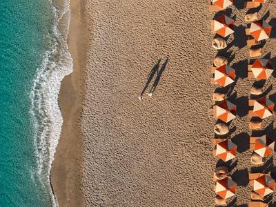 Strand mit türkisfarbenem Wasser, Sand und Schatten eines Spaziergängers neben Sonnenschirmen.