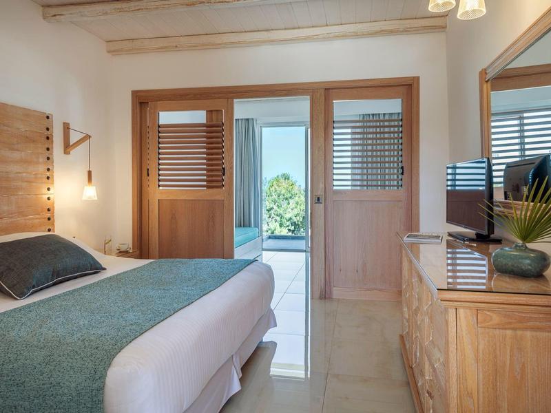 Bright hotel room with double bed, wooden furniture, and view of a terrace with garden.