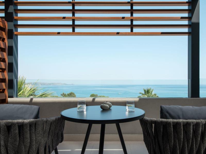 Balcony with table and two chairs overlooking the sea under a wooden slatted roof.