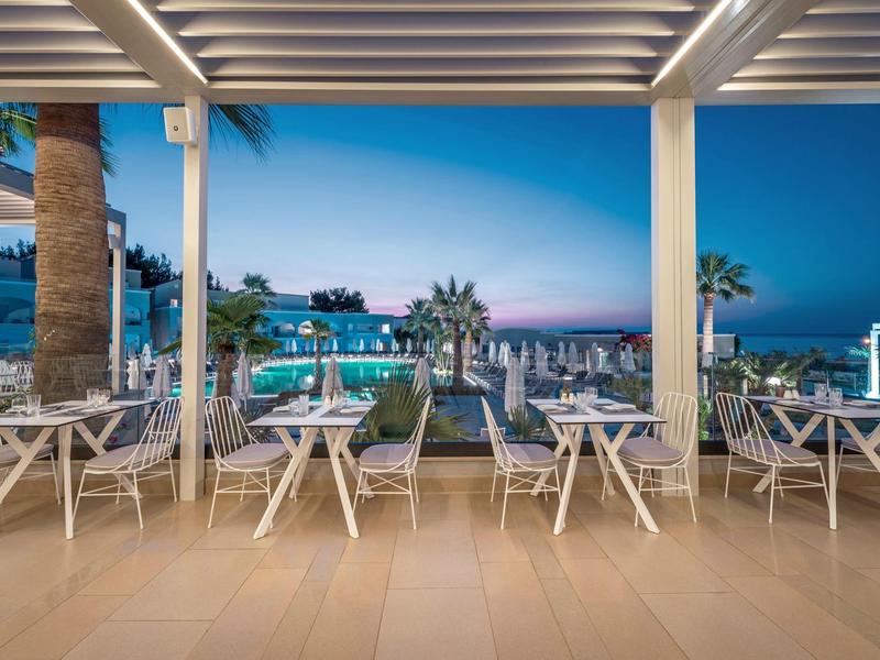 Terrace with white tables and chairs overlooking a lit pool and palm trees at dusk