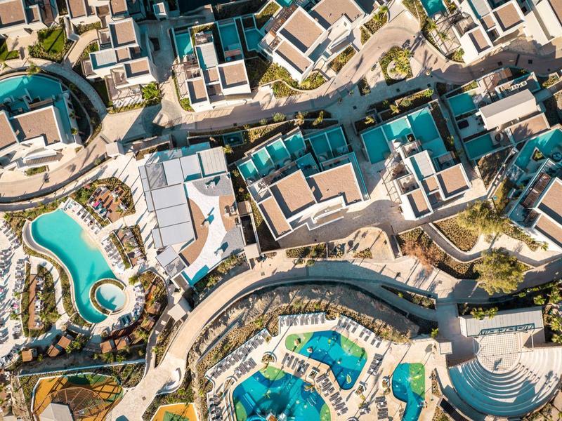 Aerial view of a resort with multiple pools, buildings, and pathways in a sunny landscape.