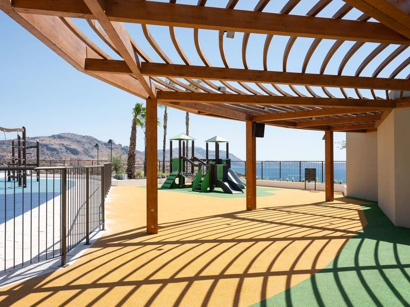 Sunny terrace with wooden pergola, view of playground and sea in the background.