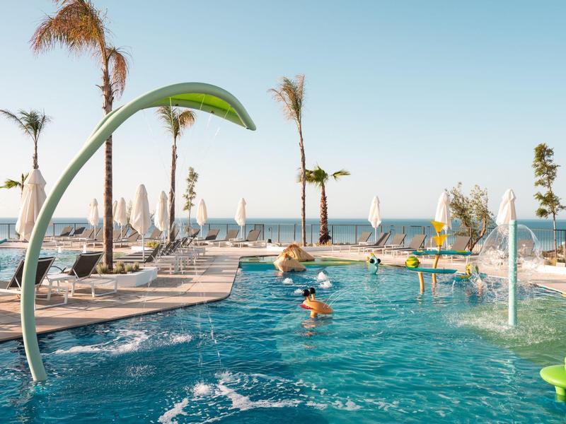 Modern pool area with sun umbrellas, palm trees, and sea view under clear sky.