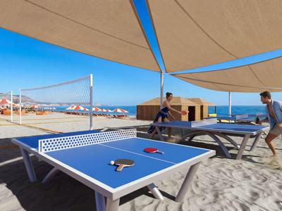 Zwei Personen spielen Tischtennis unter einem Sonnensegel am Sandstrand mit Blick aufs Meer.