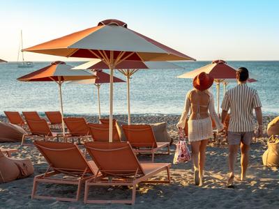 Ein Paar geht am sonnigen Strand mit Liegestühlen und Schirmen entlang.
