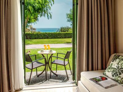 Blick aus Hotelzimmer auf Terrasse mit Tisch, zwei Stühlen und Garten bis zum Meer.
