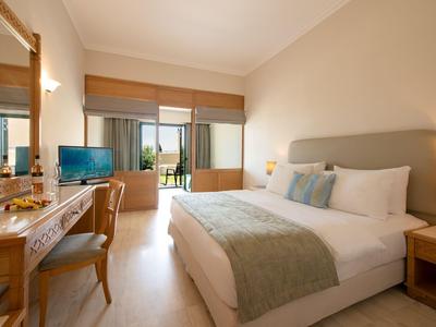 Helles Hotelzimmer mit großem Bett, Schreibtisch, Fernseher und Balkon mit Stadtblick.