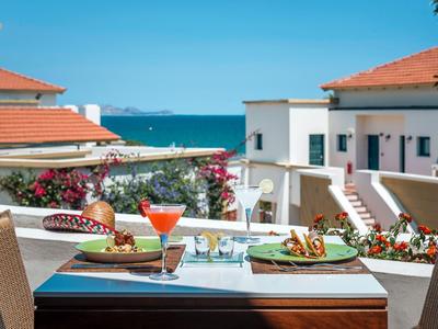 Tisch auf Balkon mit Essen, Getränken und Blick auf Hotelgebäude und Meer im Hintergrund.