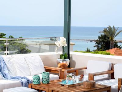 Cozy outdoor seating with wooden table overlooks ocean and greenery under clear sky.