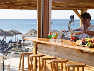Bar counter with wooden stools overlooking beach umbrellas and ocean, bartender preparing drinks.