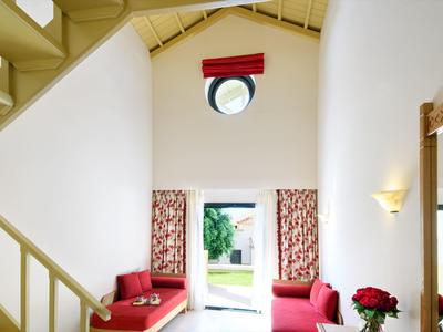 Bright hotel room with red sofas, patterned curtains, circular window, and garden view through open door.