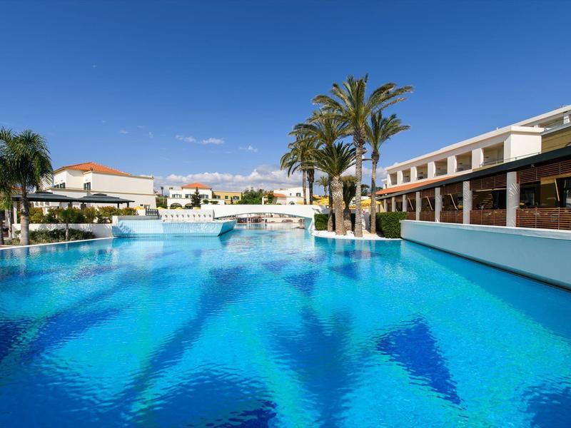 Clear blue pool with palm trees at a modern hotel under a bright sky.