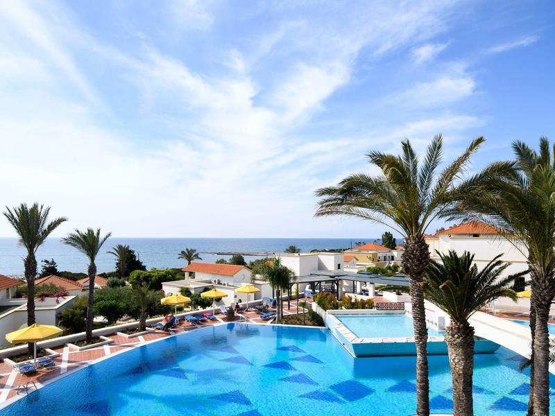 Resort pool with palm trees and sea view under a bright, partly cloudy sky.