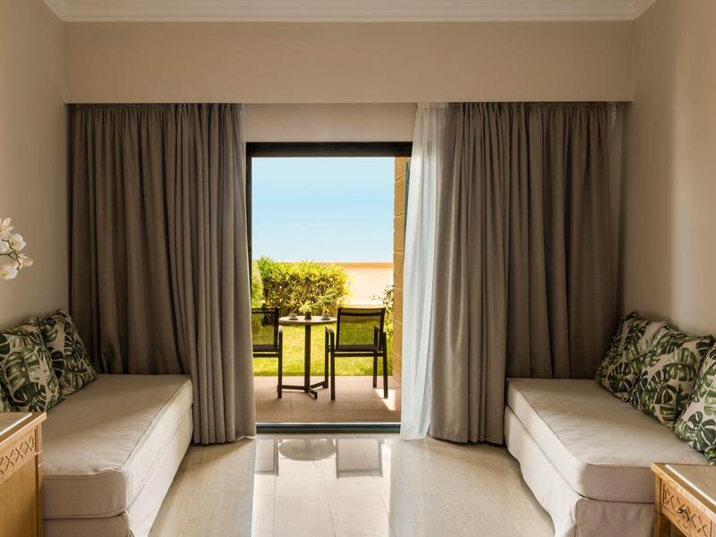 Hotel room with two sofas, floral cushions, and a balcony overlooking outdoor seating and greenery.