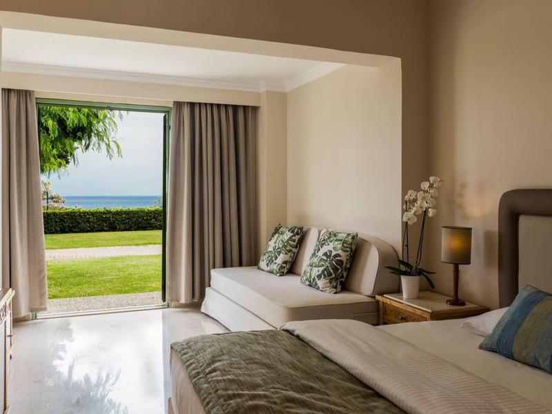 Hotel room with a bed, sofa, and open door to a green garden and ocean view.