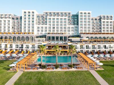 Modernes Hotel mit mehreren Etagen, großen Pools und Liegestühlen vor einer grünen Wiese.