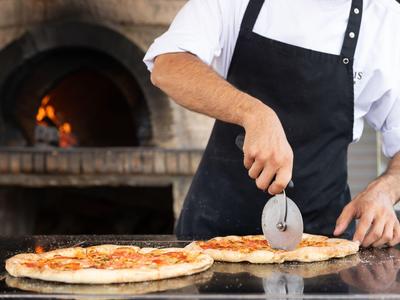 Ein Koch schneidet frisch gebackene Pizza neben einem Holzofen.
