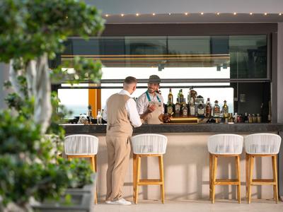 Zwei Männer stehen an einer stilvollen Hotelbar mit Hockern und Pflanzen im Vordergrund.