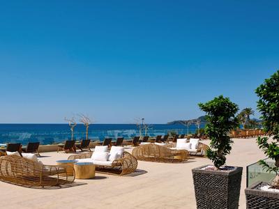 Terrazza di hotel sulla spiaggia con mobili in rattan, ombrelloni e vista sul mare sotto un cielo blu.