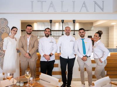Sechs Hotelmitarbeiter stehen in eleganter Arbeitskleidung in einem modernen italienischen Restaurant.