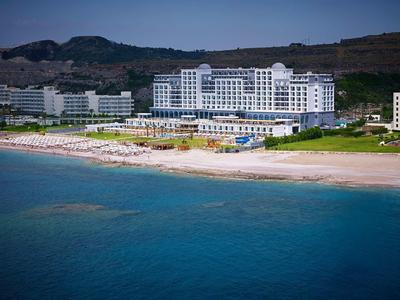 Grande hotel con spiaggia e lettini di fronte a un paesaggio collinare sotto un cielo azzurro.