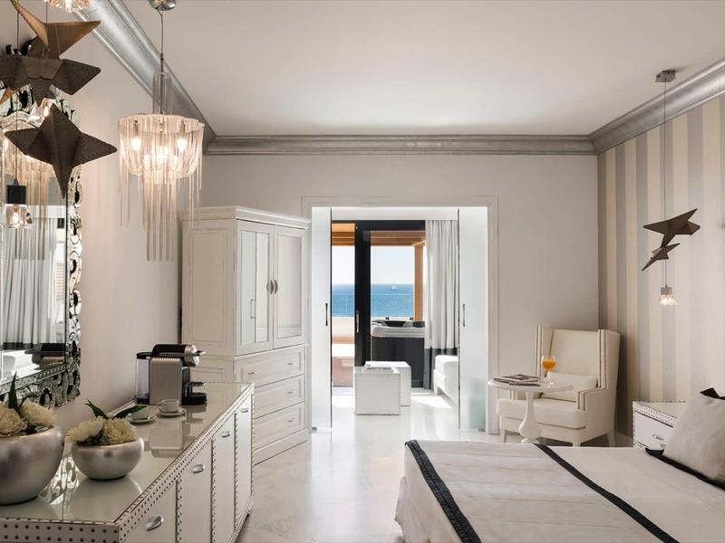 Camera d'hotel luminosa con tonalità neutre, zona salotto e vista sul mare.