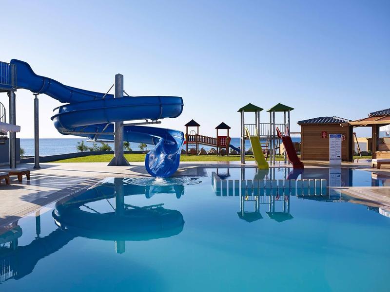 Una piscina all'aperto con uno scivolo blu e varie strutture giochi colorate sotto un cielo sereno.